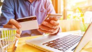 Човек, който използва кредитна карта и смартфон, докато седи пред лаптоп на маса.