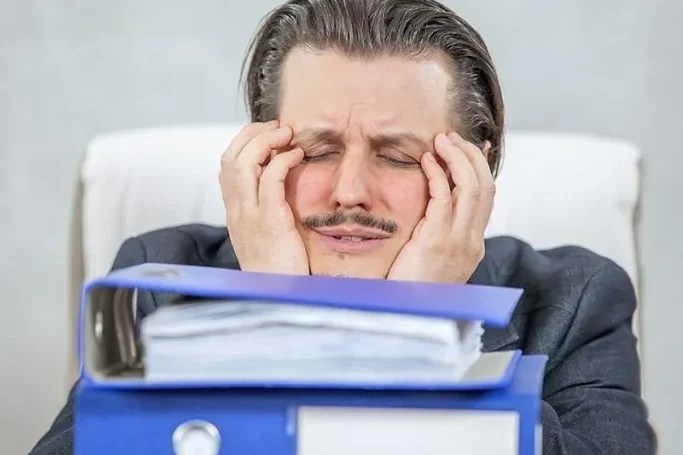 Мъж с преморен вид, държащ си лицето с ръце, пред бюро с папки и документи на фона на сива стена.